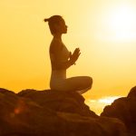 A woman meditates in a yoga pose on rocks during a warm, golden sunset.